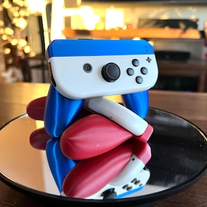 以下が含まれることがあります： 白と黒のボタンが付いた、青、白、赤の任天堂スイッチジョイコンコントローラー。コントローラーは、丸い黒色の反射面の上に置かれています