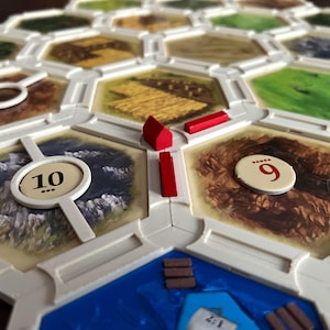 Puede incluir: Primer plano de un juego de mesa con fichas hexagonales. Las fichas son blancas con detalles marrones, verdes y azules. Hay piezas rojas en el tablero, y los números 9 y 10 son visibles.