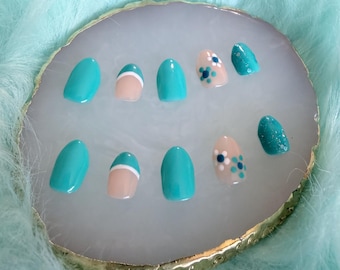 May include: A set of ten press-on nails featuring a mix of solid turquoise, turquoise French tips with white lines, and nude nails with blue and white floral accents. Some nails have a turquoise glitter finish, all arranged on a round display.
