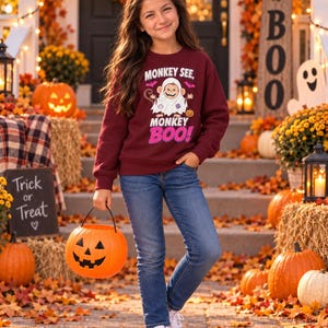 Peut inclure: Un sweat-shirt à manches longues bordeaux orné d'un motif de singe déguisé en fantôme avec le texte MONKEY SEE, MONKEY BOO!, porté avec un jean bleu et des baskets blanches, tenant un seau à bonbons en forme de citrouille orange vif.