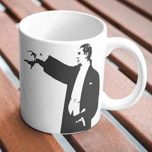 Peut inclure: Mug en céramique blanche avec une silhouette noire et blanche d'un homme en smoking, gesticulant avec sa main droite. La tasse a une anse incurvée et est posée sur une surface en bois.