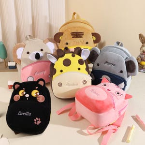 May include: A collection of plush animal-shaped backpacks in various colors. The backpacks include a koala, tiger, giraffe, elephant, cat, and a black cat. Each backpack has a name embroidered on it. The backpacks have adjustable straps and zippers.