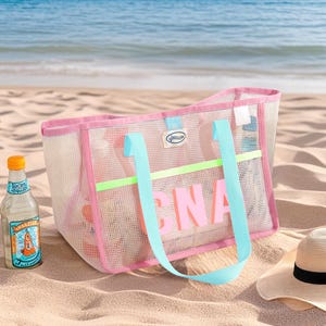 May include: A pink mesh beach tote bag with the word "PINA" in large, pink letters. The bag has light blue handles and a neon green stripe. A bottle of beverage and a straw hat are next to the bag on the sand.