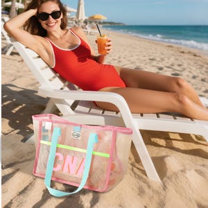 May include: A pink mesh beach tote bag with light blue handles and a neon green stripe. The bag has the letters "CNA" in pink. A woman in a red swimsuit and sunglasses is relaxing on a beach chair.