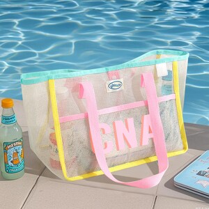 May include: A white mesh tote bag with pink handles and the letters "CNA" in pink. The bag has yellow and teal trim. A bottle of lemonade and a small notebook are next to the bag. The background is a pool of water.