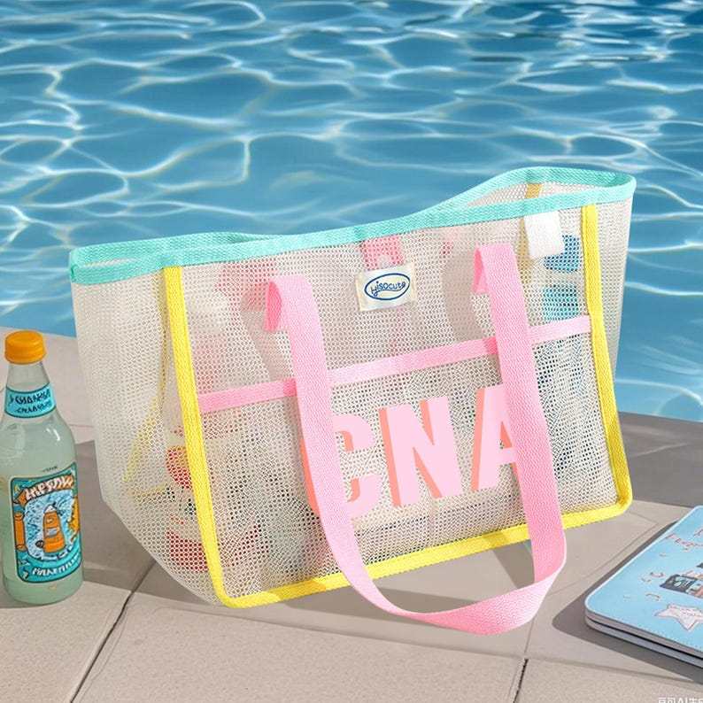May include: A white mesh tote bag with pink handles and the letters "CNA" in pink. The bag has yellow and teal trim. A bottle of lemonade and a small notebook are next to the bag. The background is a pool of water.