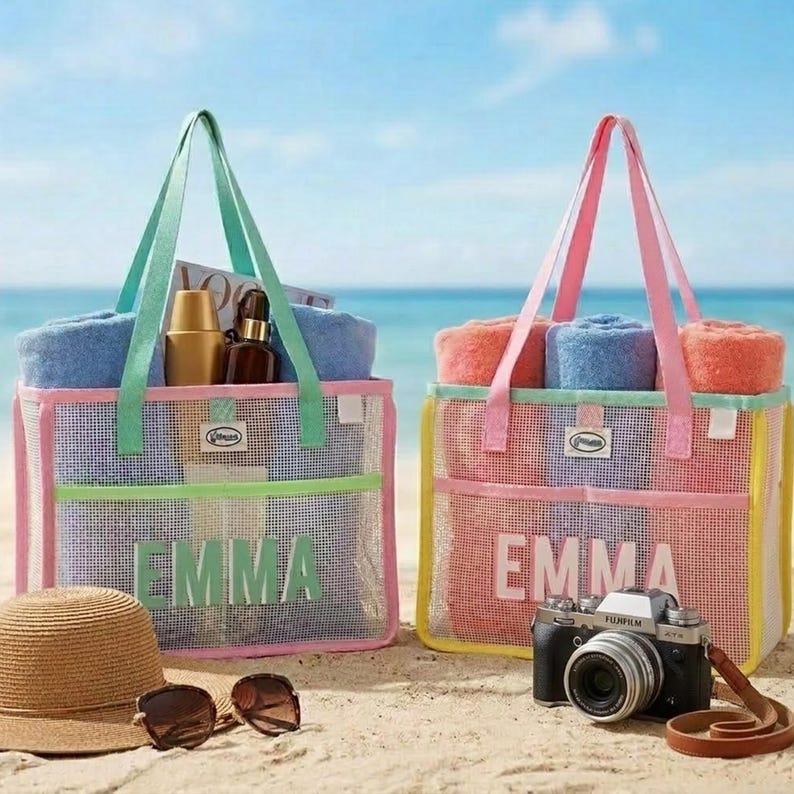 May include: Two mesh beach bags with colorful trim and handles, personalized with the name "EMMA". Each bag contains rolled towels and other beach essentials. A straw hat, sunglasses, and a camera are on the sand.
