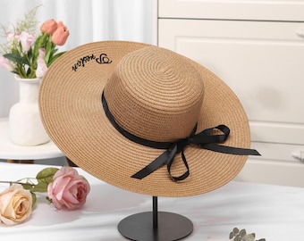 May include: A wide-brimmed, woven straw hat with a black ribbon and bow. The hat has the word "Preston" embroidered on the brim. The hat is displayed on a black metal stand, suitable for sun protection.