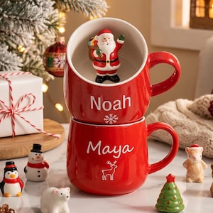 Könnte beinhalten: Zwei gestapelte rote Keramiktassen mit weißem Innenraum. Die obere Tasse enthält eine 3D-Weihnachtsmannfigur und den Namen Noah mit einer Schneeflocke. Die untere Tasse zeigt den Namen Maya und die weiße Umrisszeichnung eines Rentiers.