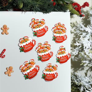 May include: A collection of Christmas magnets. The magnets depict gingerbread people in red mugs with the text "Merry Christmas". The mugs have white frosting and holly accents. Two gingerbread men and a candy cane are also present.