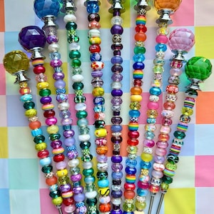 May include: A collection of decorative beaded skewers with colorful glass and crystal toppers. Each skewer features a unique combination of beads in various colors, including green, blue, purple, and orange. The toppers are faceted and come in different shapes.