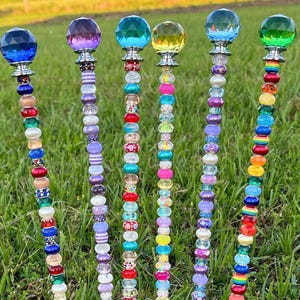 May include: Six colorful beaded garden stakes with crystal tops. The stakes are made of metal and have a variety of beads in different colors and patterns. The crystal tops are clear, blue, green, purple, yellow, and pink.