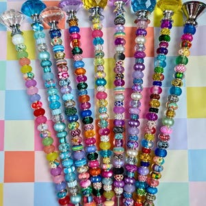 May include: A collection of colorful beaded cocktail stirrers. Each stirrer features a unique combination of beads in various colors, including blue, pink, green, and purple. The tops are adorned with decorative crystal or glass accents. The stirrers are arranged against a checkered background.