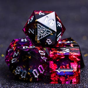 May include: A set of polyhedral dice in shades of black, purple, and red with gold flecks. The dice have white numbering and symbols. The largest die is a twenty-sided die. The dice are designed for tabletop gaming.