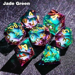 May include: A set of seven polyhedral dice in shades of jade green, teal, and purple. The dice have gold-colored numbers and are filled with gold flakes. The text "Jade Green" is in the upper left corner.
