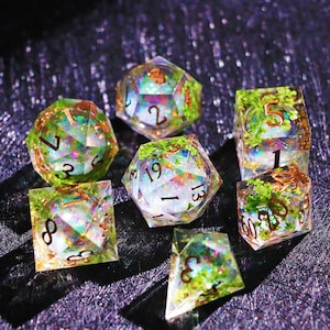 May include: Set of seven polyhedral dice with a clear resin base, containing green foliage and iridescent glitter. The dice have gold-colored numbers and are designed for tabletop role-playing games. The dice include a D4, D6, D8, D10, D12, D20, and a percentile die.