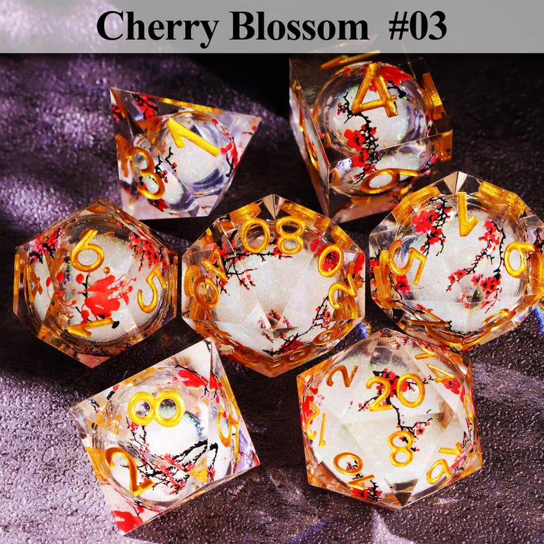 May include: Set of seven polyhedral dice with a cherry blossom design. The dice are clear with gold numbering and red and white floral inclusions. The text "Cherry Blossom #03" is at the top.