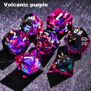 May include: Set of seven polyhedral dice in volcanic purple with white numbering and flecks of gold. The dice are a mix of purple, pink, and blue, with a translucent appearance. The text "Volcanic purple" is in the top left corner.