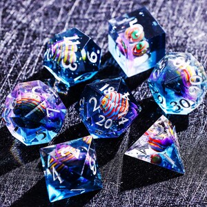 May include: A set of seven polyhedral dice with a celestial theme. Each die is clear with a blue and purple hue, and contains a colorful, spherical design. The numbers are white. The dice are arranged on a dark, textured surface.