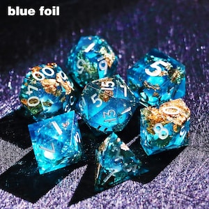 May include: Set of seven blue resin dice with gold foil inclusions. Each die has white numbering. The text "blue foil" is in the upper left corner. The dice are for tabletop games.