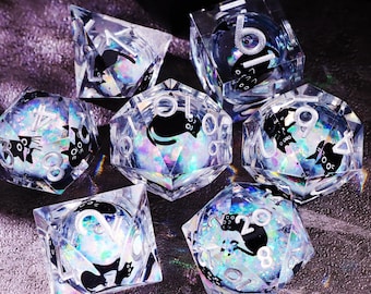Puede incluir: Un conjunto de siete dados transparentes de múltiples caras con siluetas de gatos negros y números blancos. Los dados tienen un efecto iridiscente y brillante. El conjunto incluye varias formas, como un cubo, una pirámide y una esfera. Para juegos de mesa.