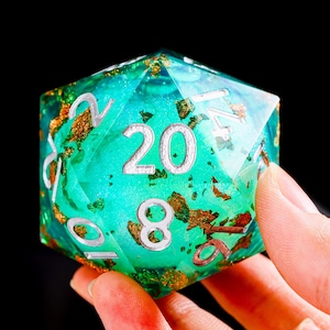 May include: A twenty-sided die made of translucent teal resin with embedded gold-coloured metallic flakes. The die features large, engraved numbers painted in a silver colour, with the number 20 prominently displayed on the front face.