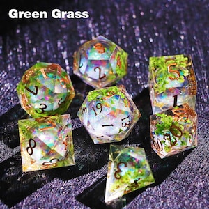 May include: Set of seven polyhedral dice with a green grass theme. The dice are clear with green and gold accents, and feature embedded greenery. The numbers are in a copper color. The text "Green Grass" is at the top of the image.