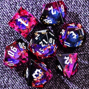 May include: A set of seven multi-sided dice for tabletop games. The dice are a mix of pink, purple, and blue with gold flecks and white numbering. The dice are arranged on a textured, dark purple surface.