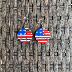 May include: A pair of patriotic earrings featuring a circular design with the American flag. The earrings display a blue field of stars, red and white stripes, and the words "God Bless America" in a script font. The earrings have silver-colored fish hook ear wires.