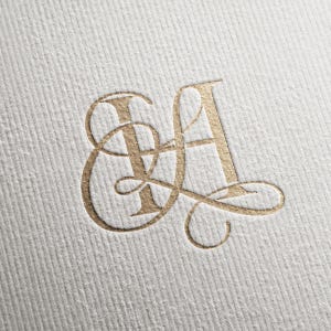 Puede incluir: Un monograma de color dorado con las letras S y A entrelazadas, representado en una tipografía serif decorativa con elegantes adornos. El diseño está grabado en relieve sobre un papel blanco texturizado con estrías diagonales.