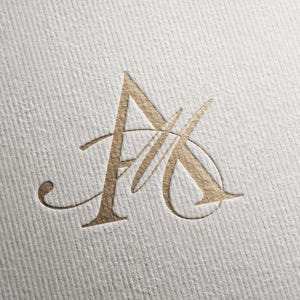 Puede incluir: Una letra A en relieve con lámina de oro metálico sobre una superficie de papel blanco texturizado. La letra presenta una fuente serif estilizada con una elegante línea curva que cruza el carácter, creando un aspecto sofisticado y dimensional.