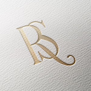 May include: A gold-colored monogram logo featuring the intertwined letters "R" and "B" in an elegant, serif font. The logo is embossed on a textured, off-white paper surface, creating a subtle shadow effect.