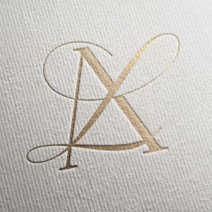 May include: A gold foil embossed monogram featuring the intertwined letters A and K on a white textured paper surface. The design uses a serif typeface with elegant, sweeping calligraphic flourishes that connect the two letters together.