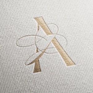 May include: A gold-colored letter "A" logo with an intricate, abstract design. The logo is embossed on a textured, off-white paper surface. The design features a stylized letter "A" with overlapping curved lines.