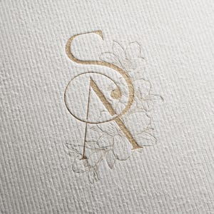 Könnte beinhalten: Ein goldfarbenes Monogramm-Design mit den verschlungenen Buchstaben "S" und "A" und floralen Akzenten. Das Design ist auf strukturiertem, cremefarbenem Papier geprägt, was einen subtilen, eleganten Effekt erzeugt. Die floralen Elemente verleihen dem Ganzen eine zarte Note.