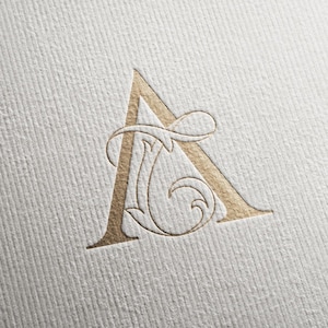 Puede incluir: Una letra A estampada en lámina de oro sobre una superficie de papel blanco texturizado. La letra presenta una tipografía con serifa con un intrincado diseño de filigrana en espiral integrado en el lado derecho, creando un efecto de monograma.