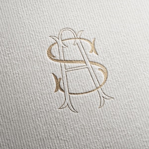May include: A gold foil embossed monogram featuring the intertwined letters S and A on a white textured paper surface. The design showcases elegant, thin lines with decorative flourishes at the terminals of each letter, creating a cohesive logo.