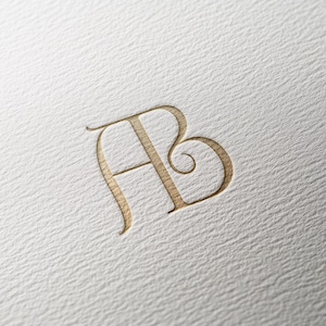 May include: A gold-colored embossed monogram featuring the letters A and B intertwined in a calligraphic script style. The design is centered on a white, textured paper surface with a visible grain, creating a subtle three-dimensional effect.