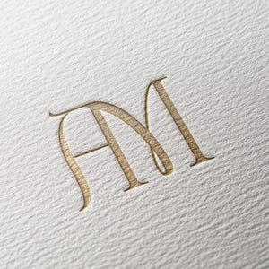 May include: Gold-colored monogram featuring the letters "A" and "M" in an elegant, flowing script. The letters are embossed on a textured, off-white paper background, creating a subtle, sophisticated design.