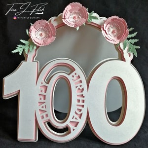 May include: White and pink layered papercraft number 100 with the word "Happy Birthday" inscribed on the left side. The number is decorated with pink paper flowers.