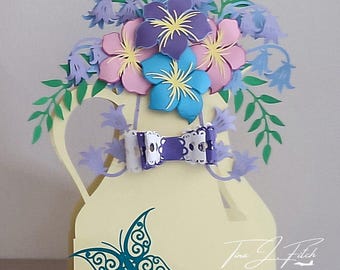 May include: A light yellow paper craft shaped like a pitcher, adorned with paper flowers in shades of purple, pink, and blue. A decorative butterfly and bow add detail. The design includes the text "Tina J Fitch" and an Instagram handle.