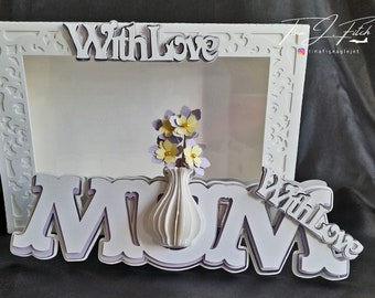 May include: White frame with a white cutout of the word "MOM" and a vase with purple and yellow flowers. The frame has a white border with a decorative pattern. The words "With Love" are written on the top and bottom of the frame.