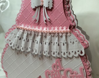Puede incluir: Una tarjeta de papel artesanal rosa y blanca con forma de vestido de bebé con un lazo blanco y las palabras "A Little Girl" en el frente.