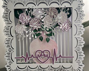May include: A white card with a decorative frame and a pink vase with a heart that says "MUM" inside. The vase is filled with white and purple flowers and has a heartbeat line below it.