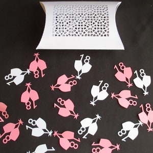 Puede incluir: Confeti de papel rosa y blanco en forma de copas de champán con el número 18. El confeti está esparcido sobre una superficie negra.