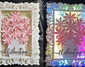 May include: Two white, intricately designed frames with a pink paper flower bouquet inside. The frame on the right has a multi-colored light shining through it. Both frames have the text "Mother's Day" written inside.