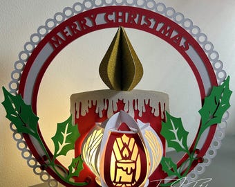 Op de afbeelding: Een rood en wit papieren knutselwerk met een kaars en een gouden ster bovenop. Het knutselwerk is versierd met groene hulstbladeren en de woorden "Merry Christmas" in een witte cirkel.