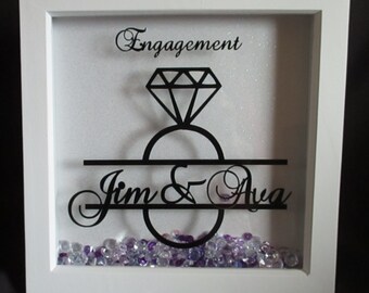 May include: White shadow box with a black silhouette of a diamond ring and the text "Engagement Jim & Ava". The box is filled with purple and clear glitter.