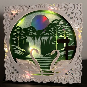 May include: A white, intricately designed frame with a circular cutout. Inside the cutout is a green, glittery scene with a waterfall, trees, and a moon. Two white swans are in the foreground, with their necks intertwined. The frame is illuminated with lights.