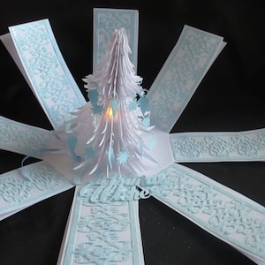 Pode incluir: Um cartão de Natal de papel branco com uma árvore de Natal pop-up e um design de padrão azul claro. O cartão está aberto e a árvore está iluminada com uma pequena luz.